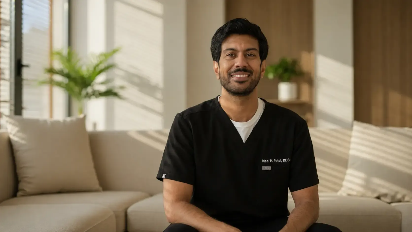 Dr. Neal H. Patel in black scrubs, seated on a sofa and smiling, in a bright room with plants and daylight through a tall window