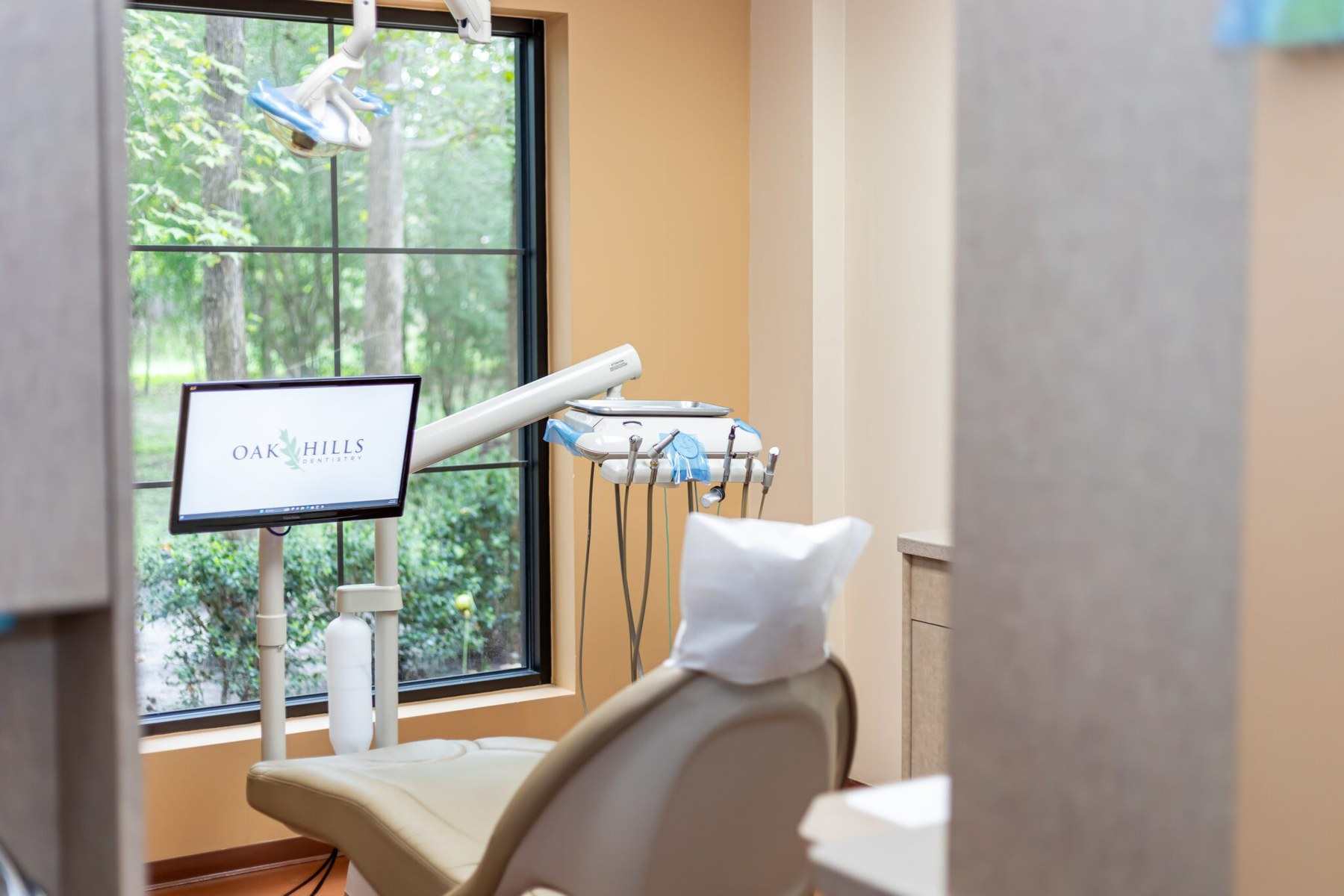 A dental chair and monitor beside a wide window filled with green trees