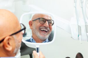 older man smiling in a hand held mirror as he observes his dental implants