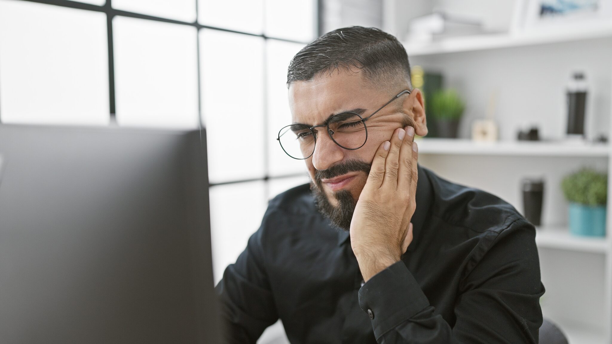 man sitting at his desk in front of computer, holding his jaw, tooth pain
